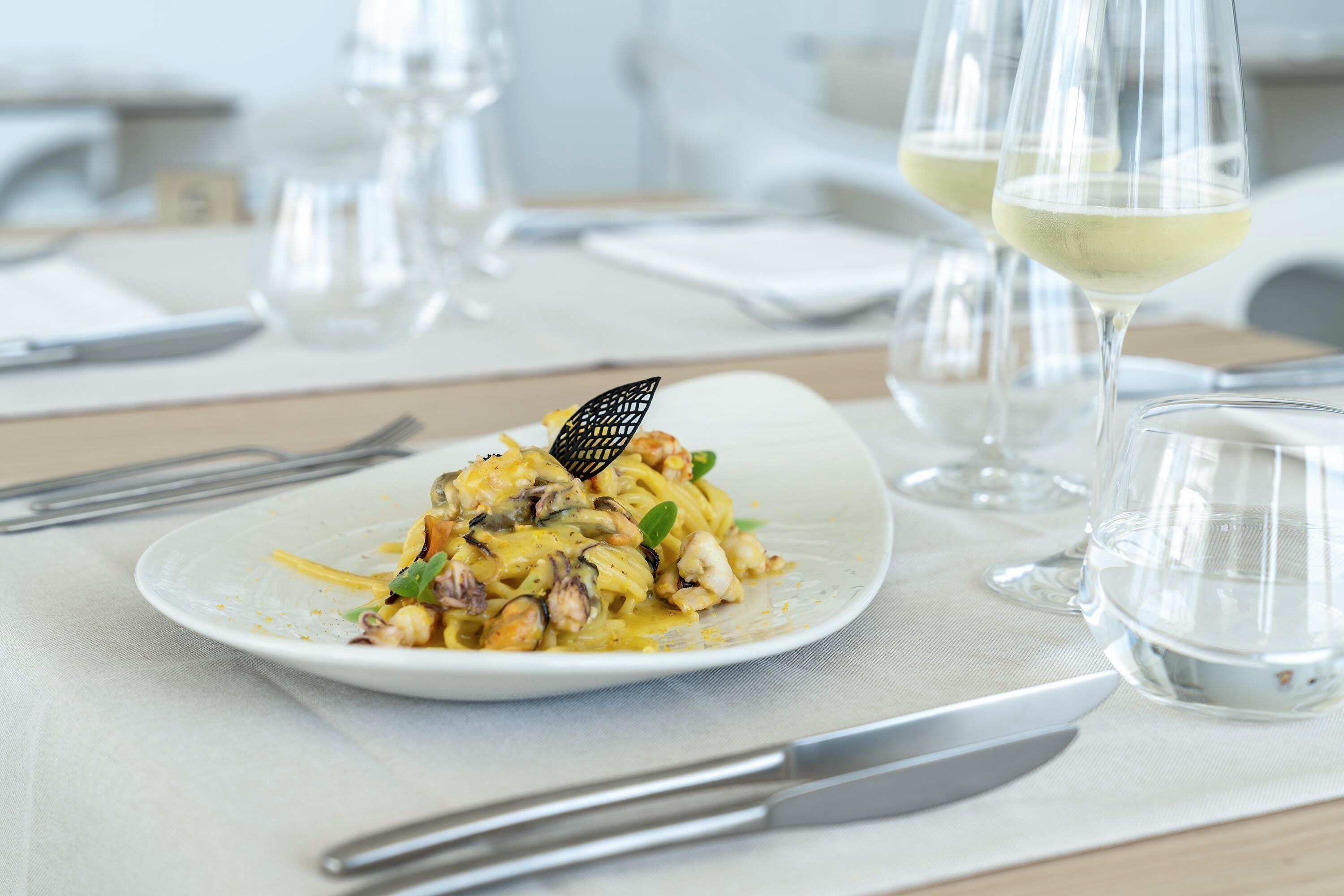 A dining table with a plate of pasta, two glasses of white wine, and cutlery.