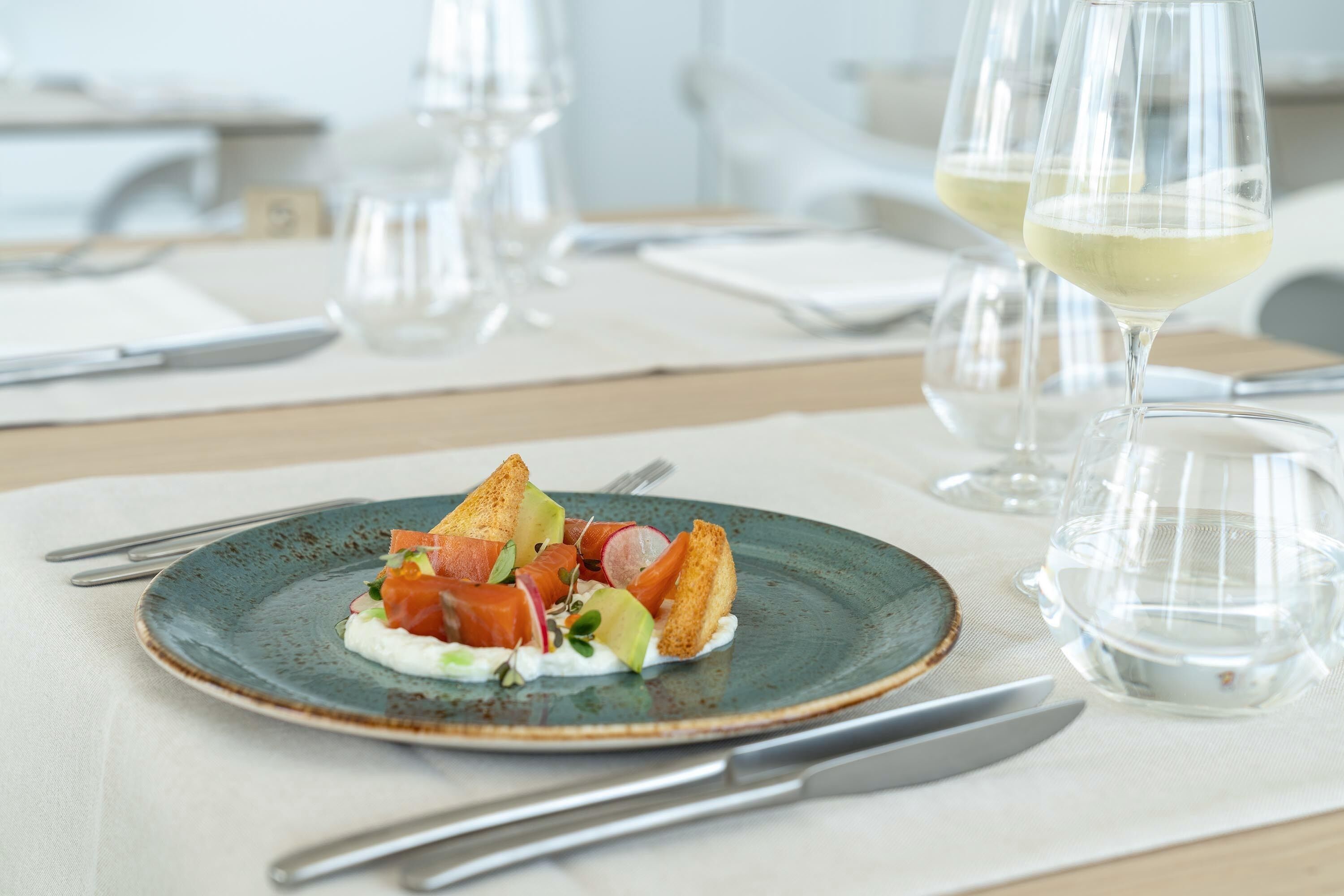 A dining table with a plate of food, two glasses of white wine, and cutlery.