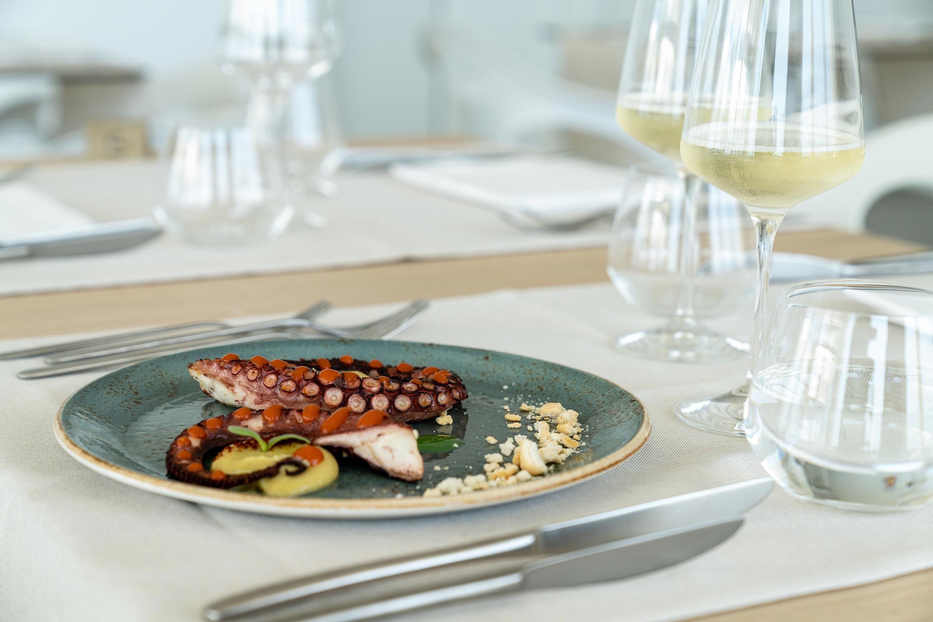 Une table à manger avec une assiette de poulpe et d’autres fruits de mer, deux verres de vin blanc et des couverts.
