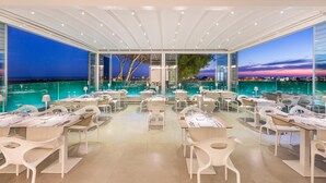 A restaurant with tables and chairs arranged around a pool, with a view of the water and a sunset in the background.