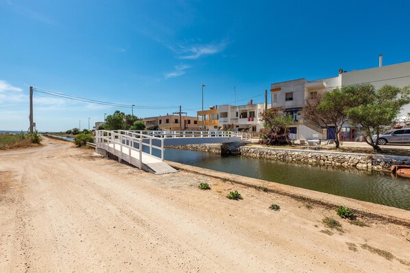 A coastal road with a white bridge over a canal, leading to a row of buildings with balconies and a clear blue sky.