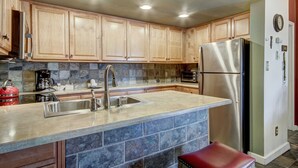 A kitchen with wooden cabinets, a stainless steel refrigerator, a sink, and a stone backsplash. - 2BR/2BA Condo on Shuttle Route - Wild Irishman 1015 - Pool & more! (Keystone)