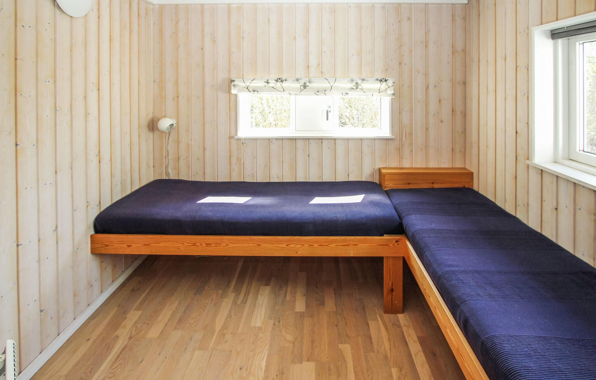 A small room with a wooden floor, a bed with a dark blue mattress, and a window with a floral curtain.