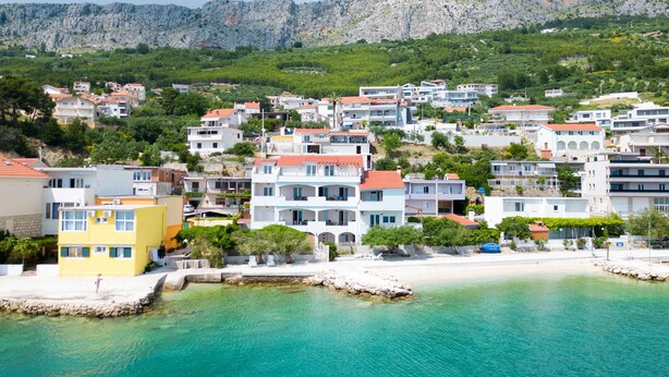 Une ville côtière avec des bâtiments le long du front de mer, une colline rocheuse en arrière-plan et des eaux turquoise et limpides.