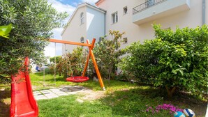 Un espace de jeux pour enfants avec une balançoire et un toboggan rouge, entouré de verdure et avec un bâtiment en arrière-plan.