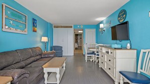 A living room with a brown leather sofa, a wooden coffee table, a blue armchair, a white dresser, and a television on a stand. - Beautiful Oceanfront Condo, breathtaking sunrises and a relaxing experience (Myrtle Beach)