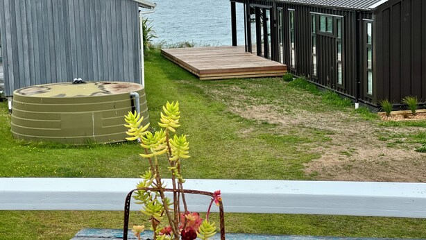 A modern house with a deck overlooking a body of water, a small garden with succulents, and a wooden bench. - Iconic 70's Kiwi Bach at Simpsons Beach  (Wharekaho, Simpsons Beach, Whitianga)