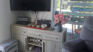 A living room with a flat-screen TV mounted on the wall, a small cabinet with drawers, a red tray with decorative items, and a window with a view of a house and greenery. - Iconic 70's Kiwi Bach at Simpsons Beach  (Wharekaho, Simpsons Beach, Whitianga)