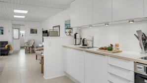 A modern kitchen with white cabinetry, a stainless steel sink, and a built-in oven. There is a dining area with a gray sofa and a wooden chair. - Awesome home in Pandrup with WiFi (Pandrup)
