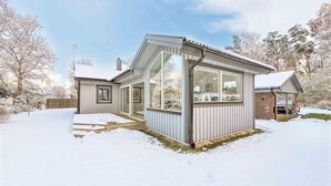 Ett modernt hus med stora fönster och en inglasad veranda, omgivet av snötäckta träd.