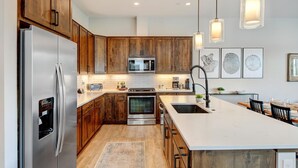 A modern kitchen with wooden cabinets, stainless steel appliances, a central island, pendant lighting, and artwork on the walls.