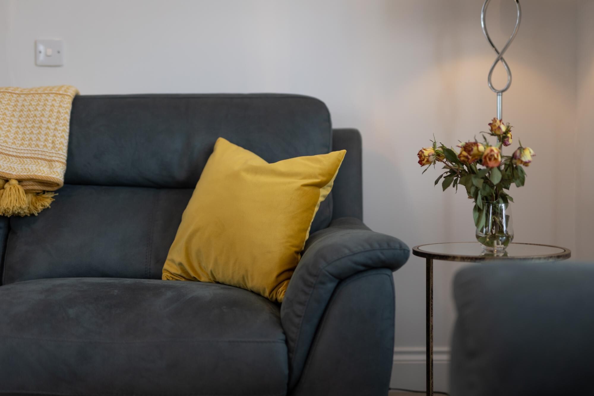 A dark blue sofa with a mustard yellow pillow and a throw blanket. A small round table with a vase of flowers is to the right.
