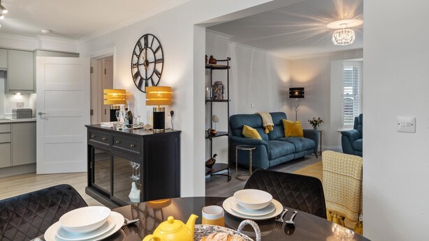 A modern living room with a blue sofa, a dining table set with plates and a yellow teapot, and a kitchen area with a black cabinet and a clock. - Cottage Mews - Seaside Cottage Parking Terrace (Tenby)