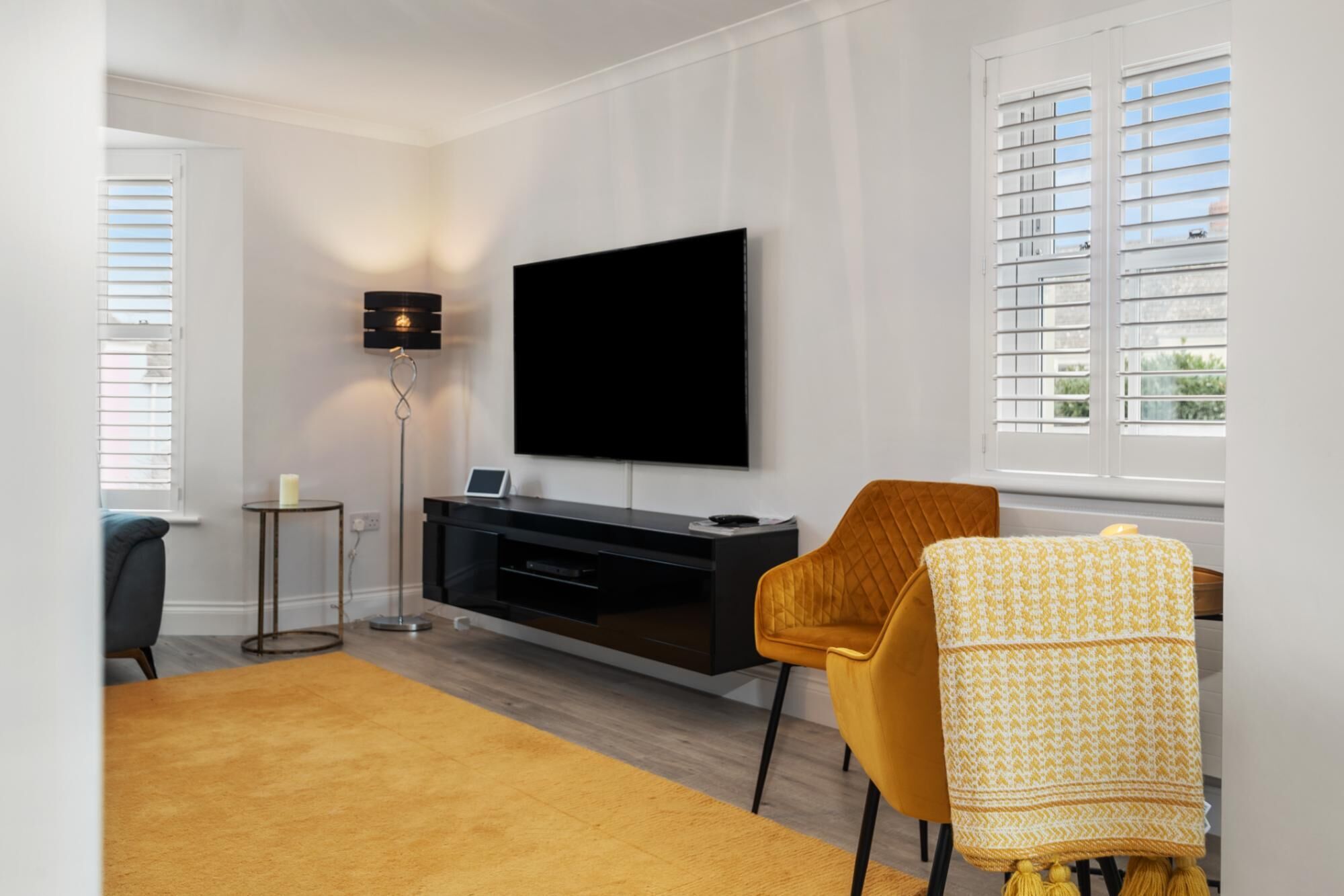 A modern living room with a flat-screen TV mounted on the wall, a black entertainment unit, a yellow armchair, and a patterned chair.