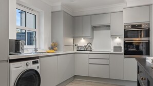 A modern kitchen with a built-in washing machine, stainless steel appliances, and a window with a view of the outside. - Cottage Mews - Seaside Cottage Parking Terrace (Tenby)