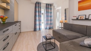 A modern living room with a grey sofa, a small black side table, and a grey rug. There is a wooden floor, a kitchen area with a counter and drawers, and a window with curtains. - Amazing apartment in Pakostane with WiFi (Pakostane)