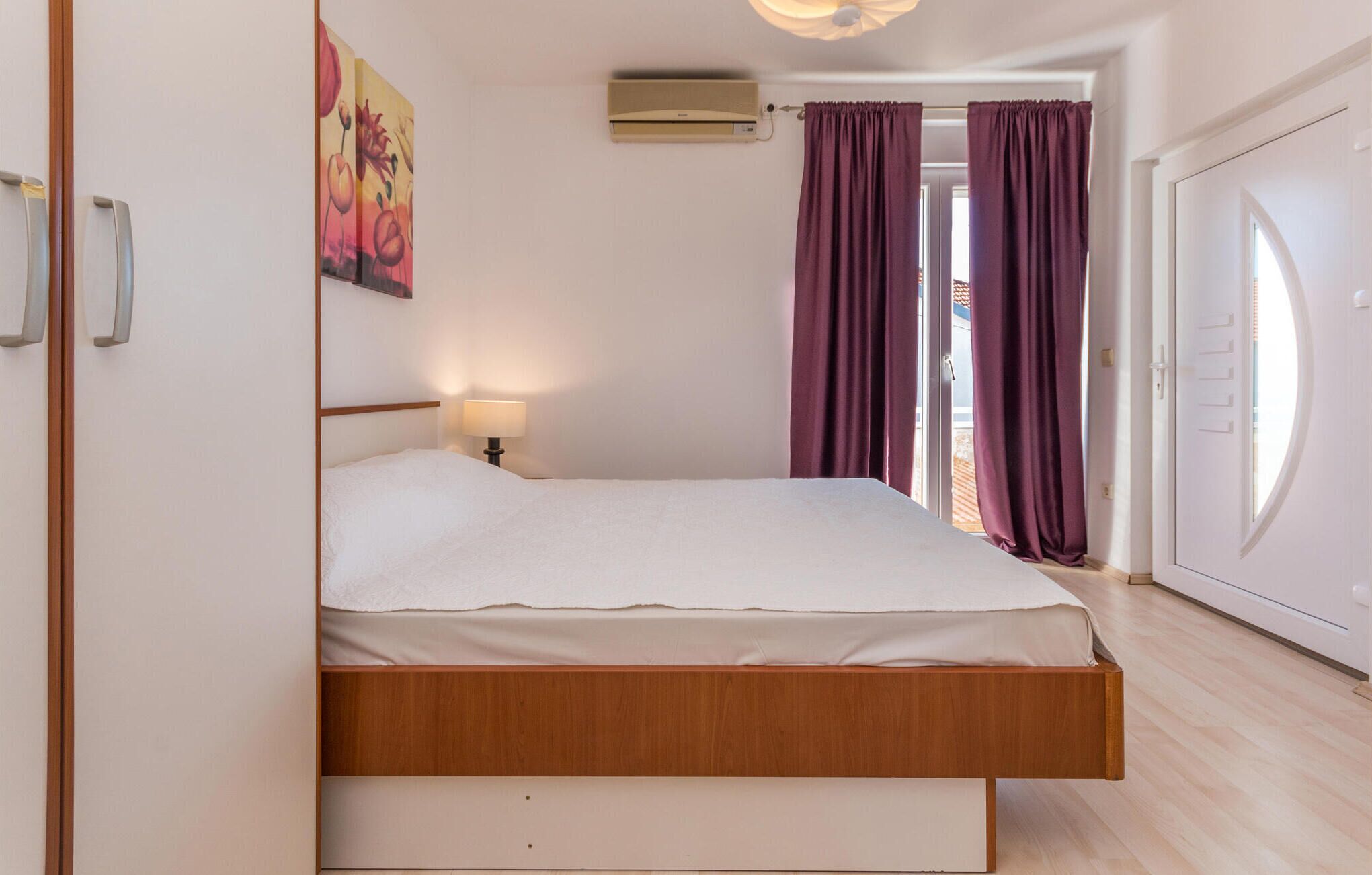 A bedroom with a bed, a nightstand, a wall-mounted air conditioner, and a door with a view to the outside.