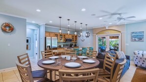 A dining area with a round table set for six, surrounded by wooden chairs. A kitchen with wooden cabinets and a dining table is visible in the background. - Private Pool! Golf Cart! Bikes! 143 Beth Lane! (Santa Rosa Beach)