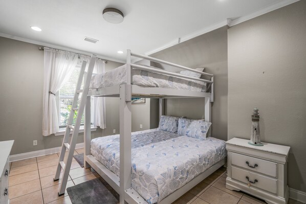 Un letto a castello con copripiumino decorato, un comodino con cassetti, una scala e una finestra con tende.