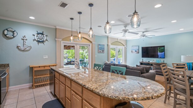 A kitchen with granite countertops, wooden cabinets, and a central island. There is a dining area with a table and chairs, a ceiling fan, and a television. - Private Pool! Golf Cart! Bikes! 143 Beth Lane! (Santa Rosa Beach)