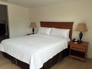 A neatly made bed with a white quilt, two bedside lamps, a wooden nightstand with a phone and a small drawer, and a wooden headboard. - One Bedroom Suite Cottage (Carmel-by-the-Sea)
