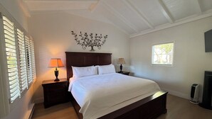 A bedroom with a large bed, two bedside tables with lamps, a decorative wall piece, a window with blinds, and a radiator. - One Bedroom Suite Cottage (Carmel-by-the-Sea)