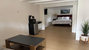 A hotel room with a kitchenette, a bed, a bedside table, a lamp, and a plant.