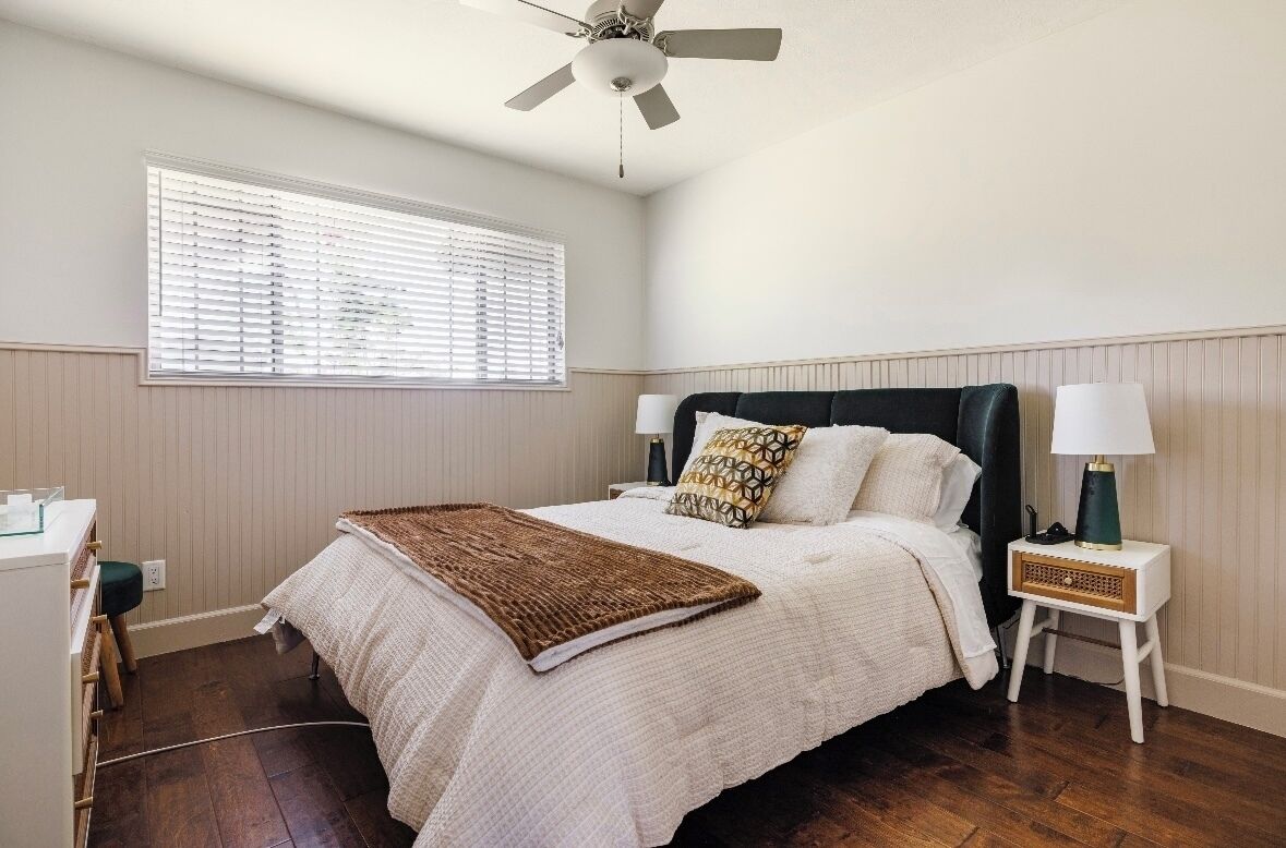 Un dormitorio con cama, ventilador de techo, mesita de noche con lámpara y ventana con persianas.
