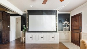 Ein Wohnzimmer mit einem großen Flachbildfernseher, der an einem weißen Schrank befestigt ist, einem Deckenventilator und einem dekorativen Baum an der Wand.