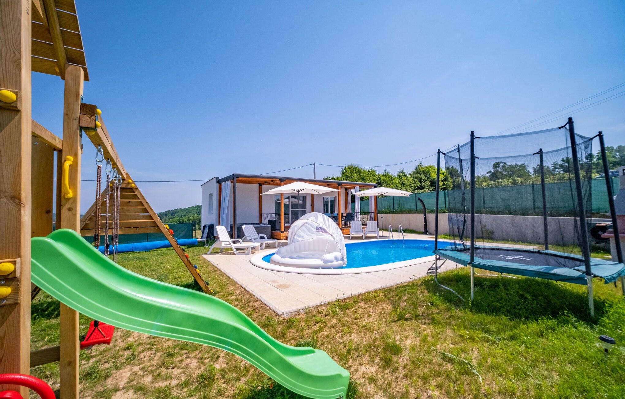 Un patio trasero con un parque infantil de madera, un tobogán verde, una piscina pequeña y un trampolín.
