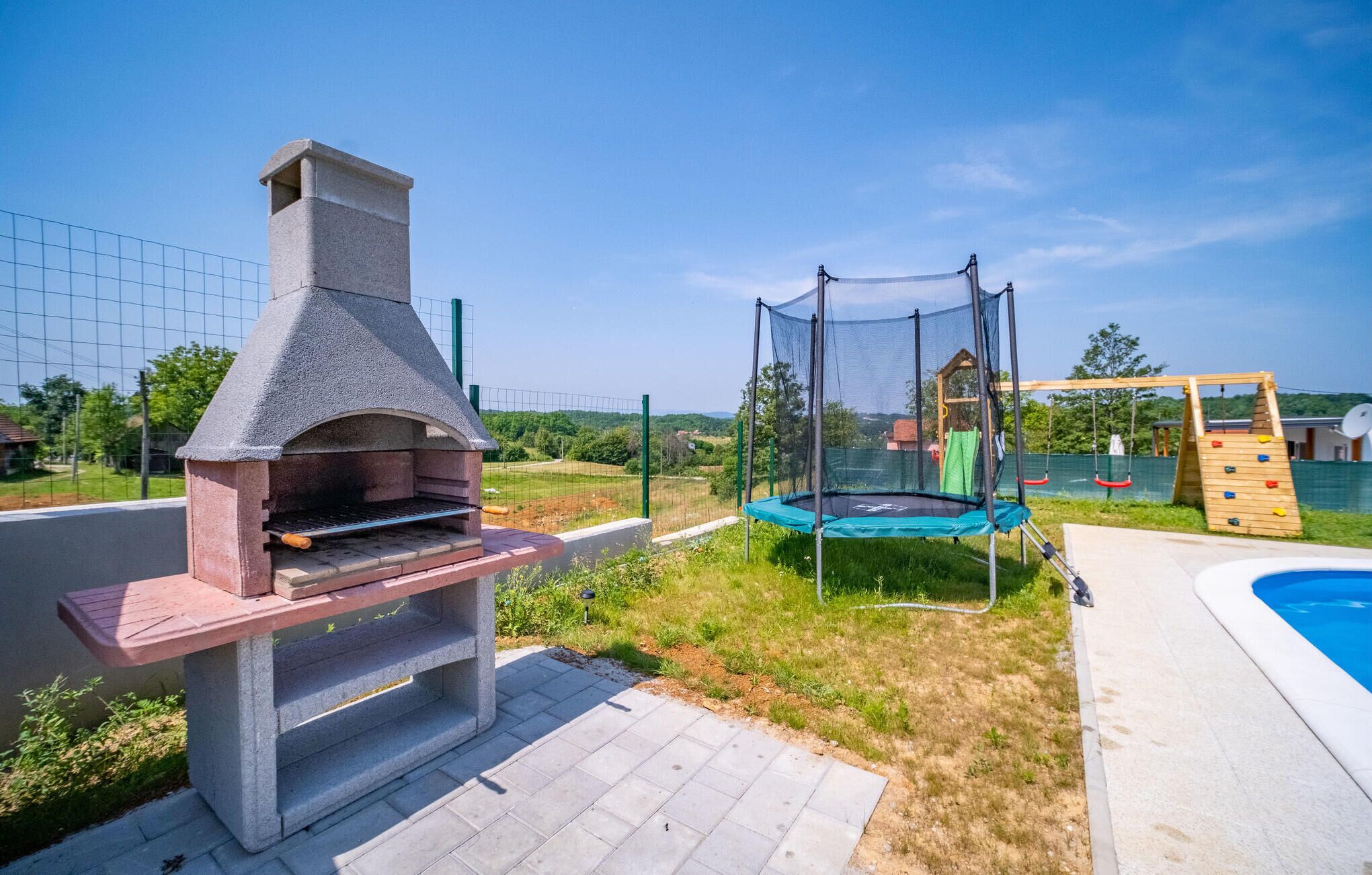 Ein Garten mit Grill aus Naturstein, Trampolin mit Sicherheitsnetz und einer hölzernen Spielanlage.