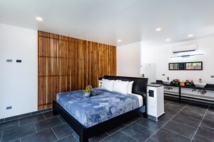 Un dormitorio moderno con cama negra, paredes blancas, un panel de madera y un baño con encimera negra y lavamanos blanco.