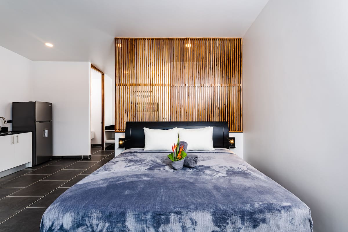 Habitación de hotel moderna con una cama grande, un revestimiento de pared de listones de madera, una pequeña área de cocina y una mesita de noche con lámpara.