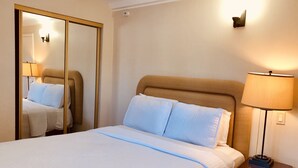 Un lit bien fait, recouvert d’une courtepointe blanche matelassée, avec deux oreillers, une lampe de chevet et un miroir posé sur une table de chevet en bois.