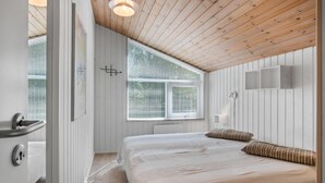 Un dormitorio con techo de madera, un ventanal con persianas, una cama con dos almohadas y una mesita de noche con lámpara.