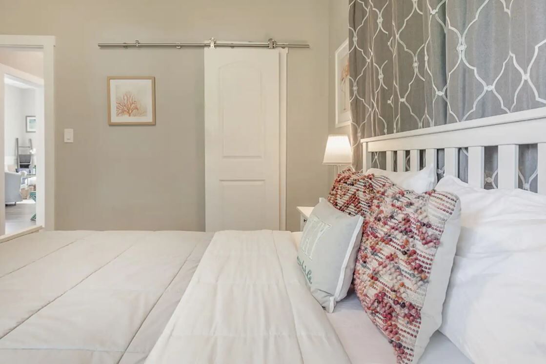 Una cama bien hecha con sábanas blancas y una manta de colores, cabecera blanca y una barra de cortina fijada a la pared.