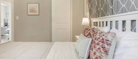 Una cama bien hecha con sábanas blancas y una manta de colores, cabecera blanca y una barra de cortina fijada a la pared.