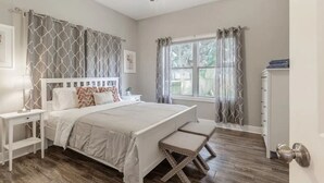 Une chambre à coucher comprenant un lit, des tables de chevet, un banc, un ventilateur de plafond et une fenêtre avec des rideaux.