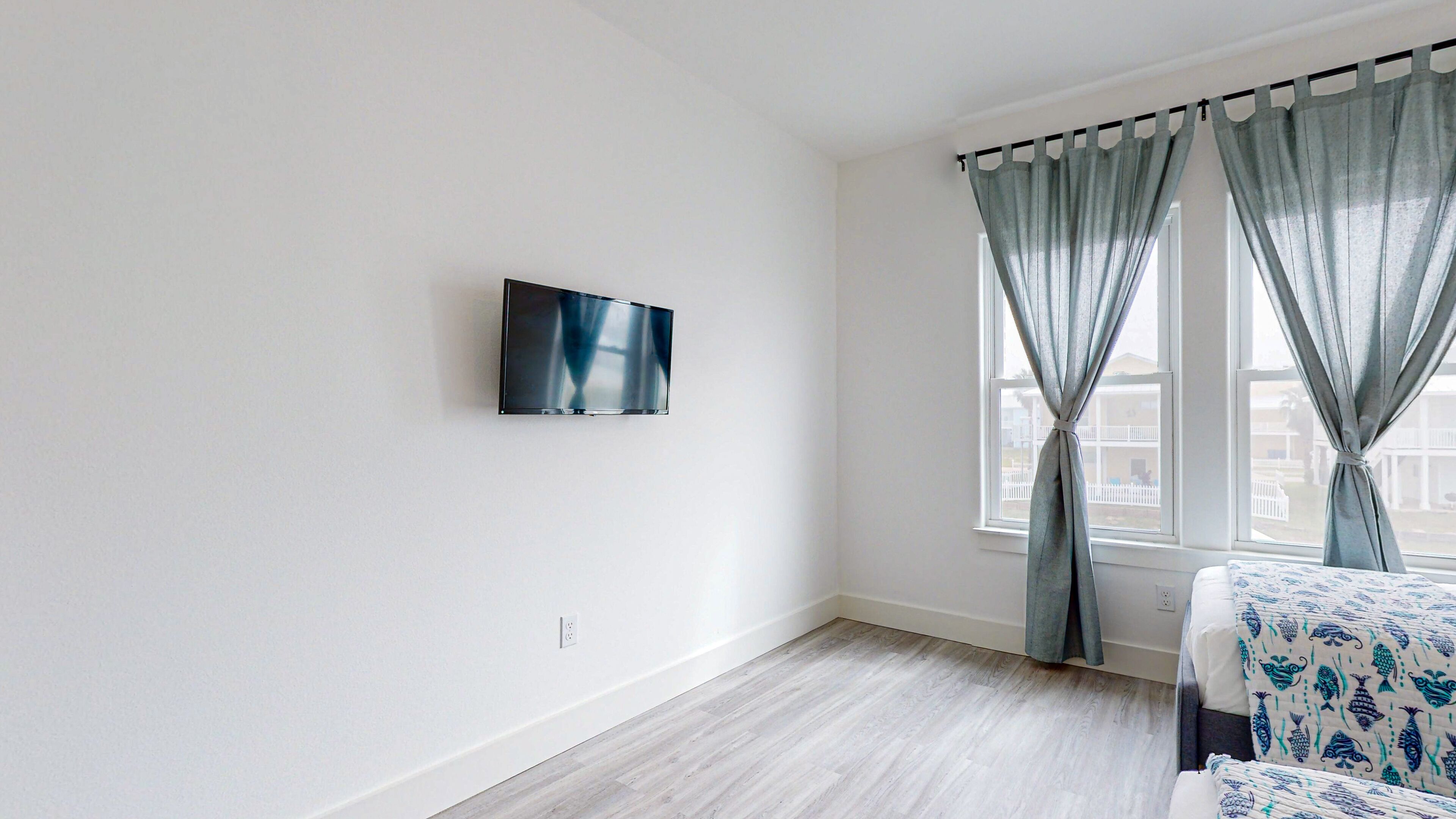 A room with a flat-screen TV mounted on the wall, a window with curtains, and a bed with patterned bedding.