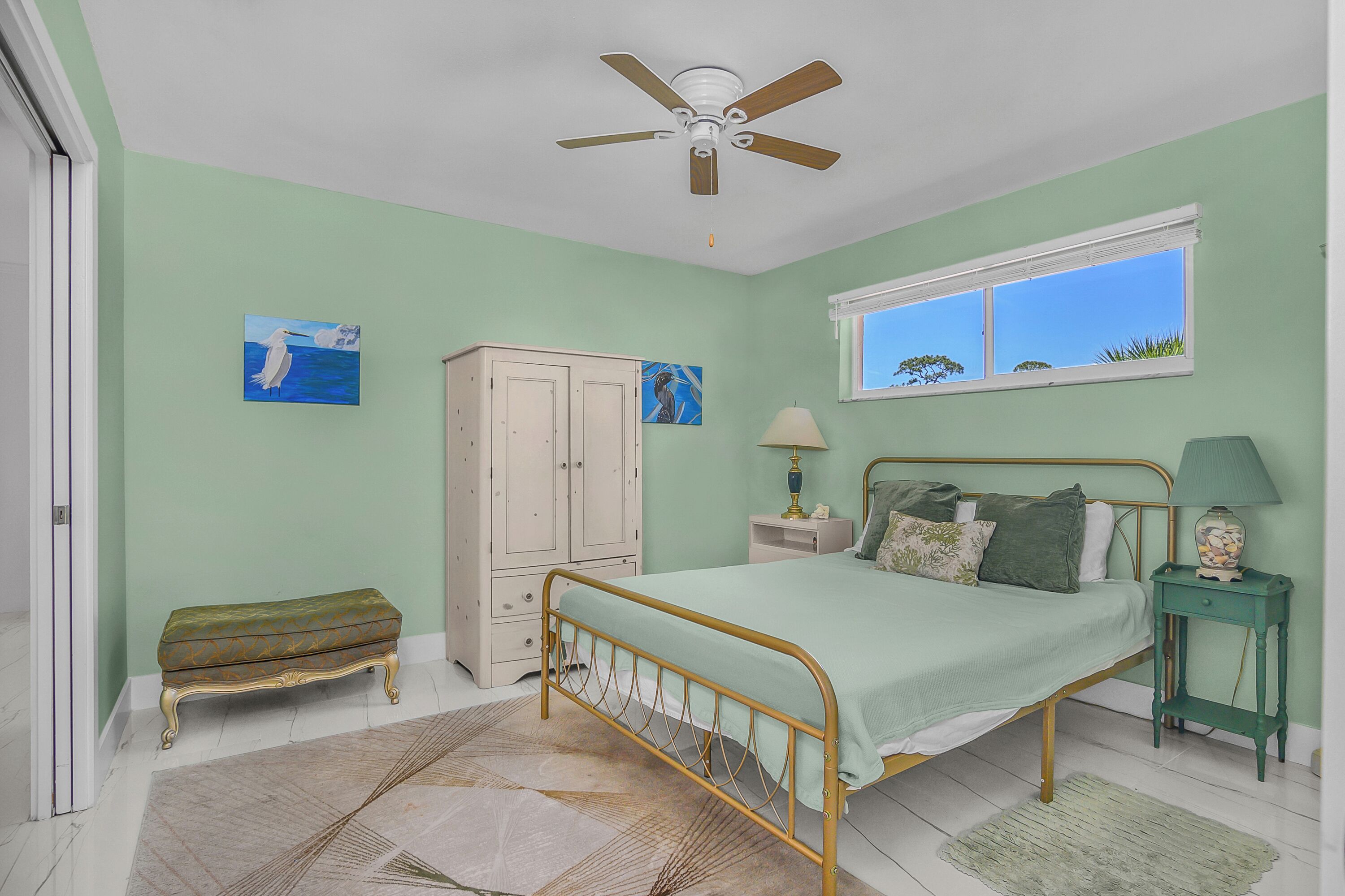 A bedroom with a bed, a ceiling fan, a window, a nightstand, a lamp, a cabinet, and a bench.