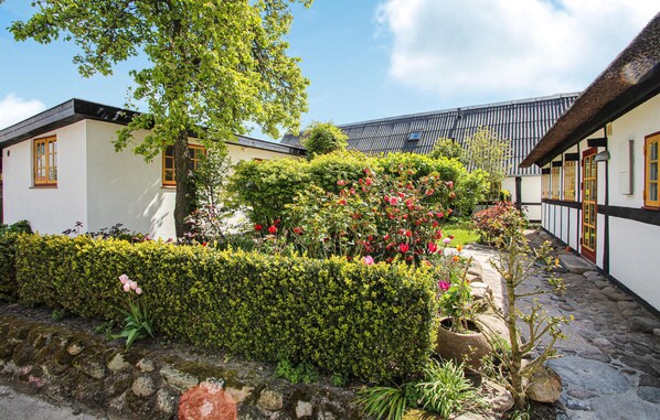 Un bâtiment traditionnel, doté d’un toit de chaume et de murs blancs, entouré d’un jardin bien entretenu où poussent diverses plantes et fleurs.