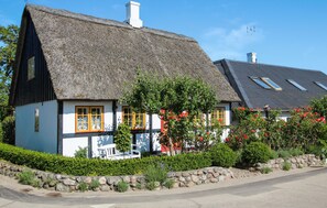 Une maison traditionnelle à toit de chaume, aux murs blancs et aux fenêtres à cadre jaune, entourée de verdure et d’un mur de pierre.
