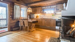 Una cocina acogedora de estilo cabaña con suelo de madera, zona de comedor con sillas, chimenea y ventana con cortinas.
