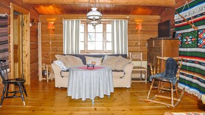 Interior de una cabaña de troncos con un sofá, una mecedora, una mesa y un televisor.