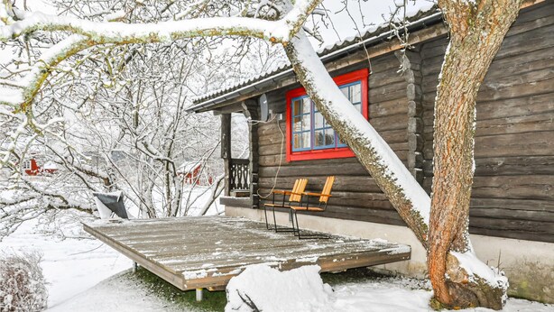 Una cabaña de madera con una ventana de marco rojo, un banco afuera y un entorno cubierto de nieve.