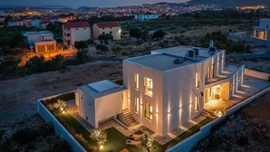 Uma casa moderna de dois andares com telhado plano, amplas janelas e área externa bem iluminada, com piscina e espreguiçadeiras.