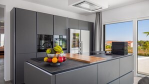 Uma cozinha moderna com armários cinza, uma ilha central com uma fruteira e vista para o exterior através de grandes janelas.