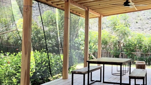 Een houten pergola met een tafel en banken, uitkijkend op een tuin met bomen en struiken.