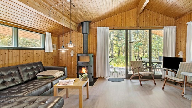 Interior acollidor d'una cabana amb un sofà de cuir, una taula de fusta, una llar de foc i una finestra gran amb vista a un bosc.