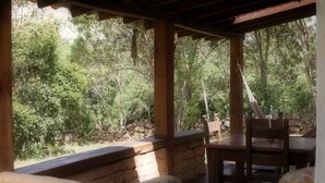 Un espacio de estar al aire libre cubierto, con paredes de piedra y muebles de madera, rodeado de abundante vegetación y árboles.